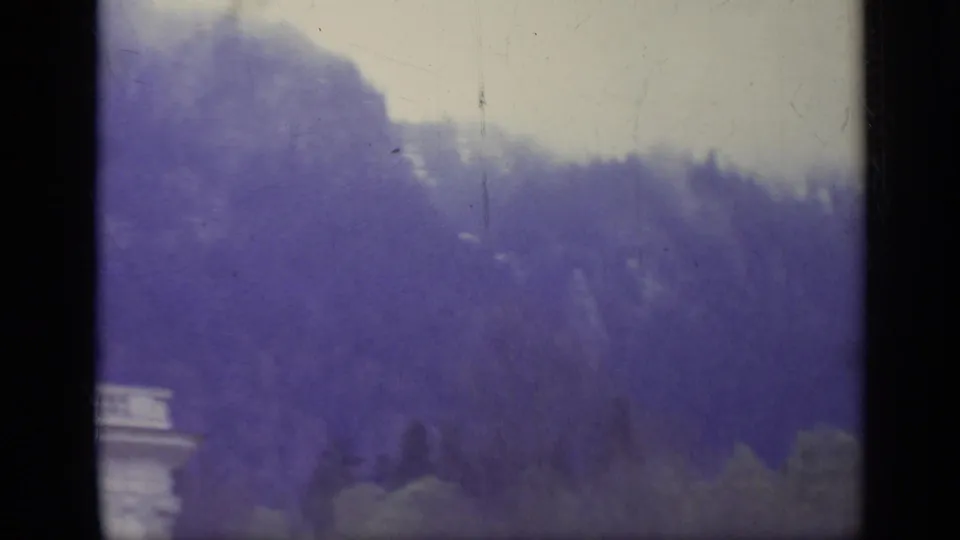 1962 Italy 8mm frame shifting away from the palace toward a wooded mountain slope, with haze, trees, cloudy sky, and a small facade edge visible.