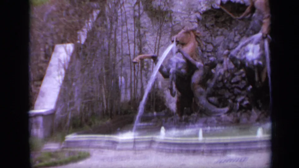 1962 Italy 8mm frame showing the fountain from the side, with a water jet, stone basin, rock wall, and garden branches.