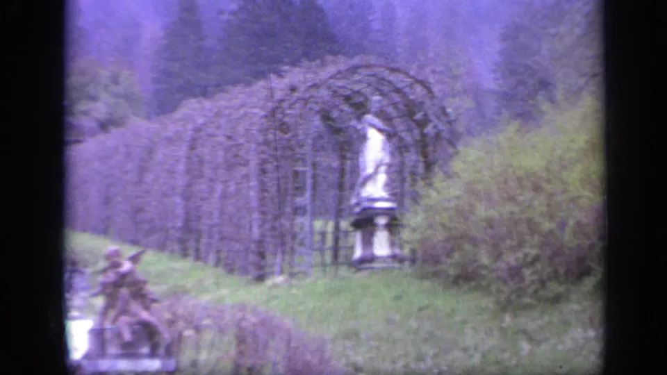 1962 Italy 8mm frame with an arched trellis tunnel, a classical statue-like figure, lawn, shrubs, and wooded hillside.