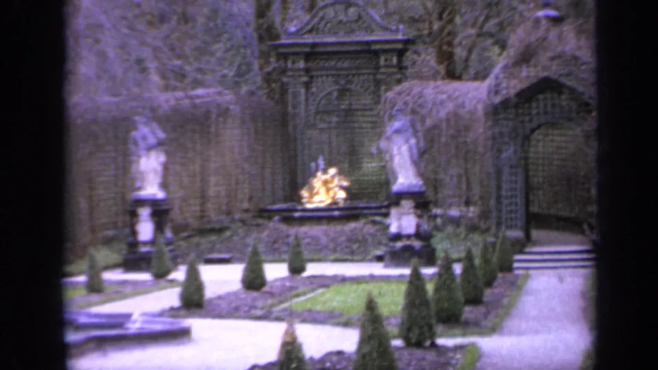 1962 Italy 8mm frame showing stone figures, clipped hedges, gravel paths, and a small gold-colored garden ornament beyond them.
