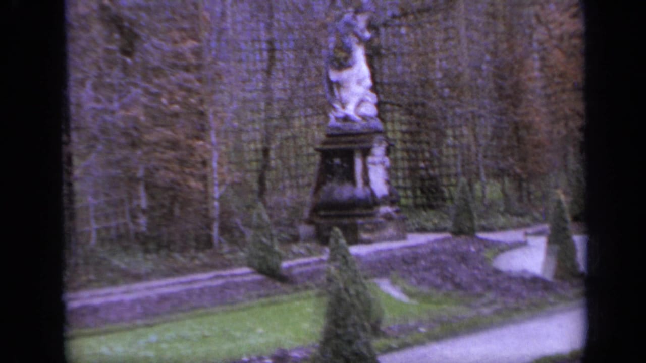 Stone pedestal statue beside lattice fencing, clipped shrubs, and curving garden paths.