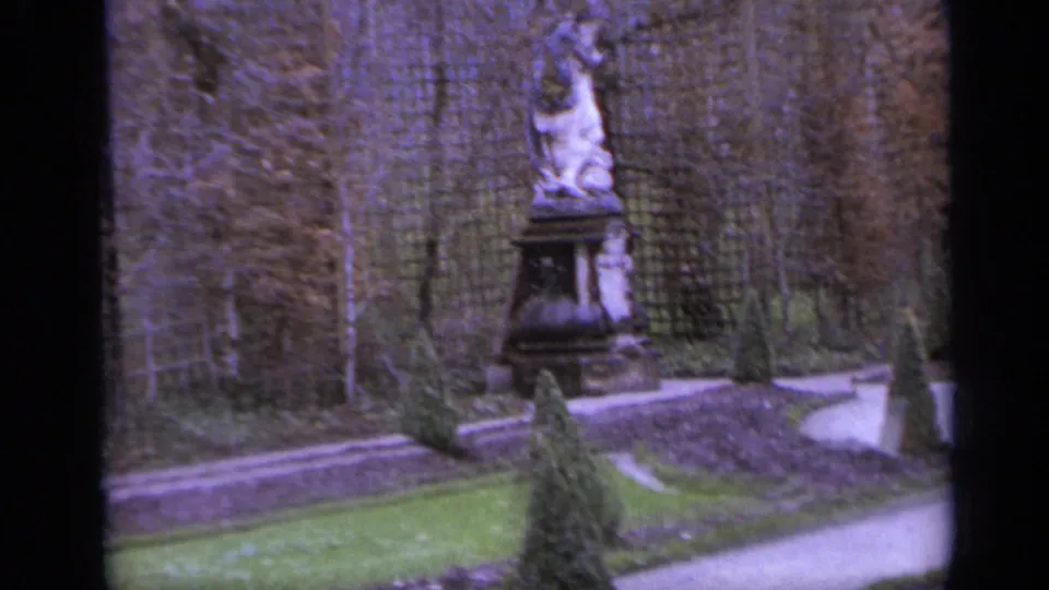 1962 Italy 8mm frame with a pedestal statue near lattice fencing, bare vines, shaped shrubs, and a curving formal garden path.