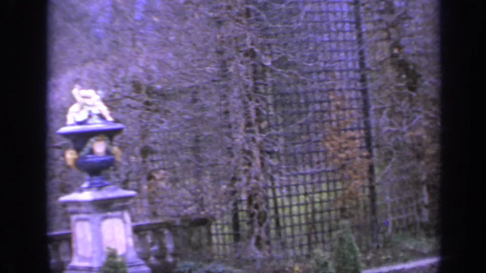 1962 Italy 8mm frame showing a decorative pedestal with a small statue, lattice fencing, bare vines, and stone garden edging.