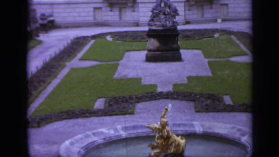 1962 Italy 8mm frame showing a formal courtyard, central sculptural monument, clipped lawns, stone paths, and a golden fountain figure.
