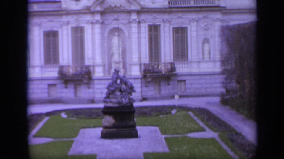 1962 Italy 8mm frame of an ornate exterior facade with balconies, a central statue niche, and a courtyard monument below.