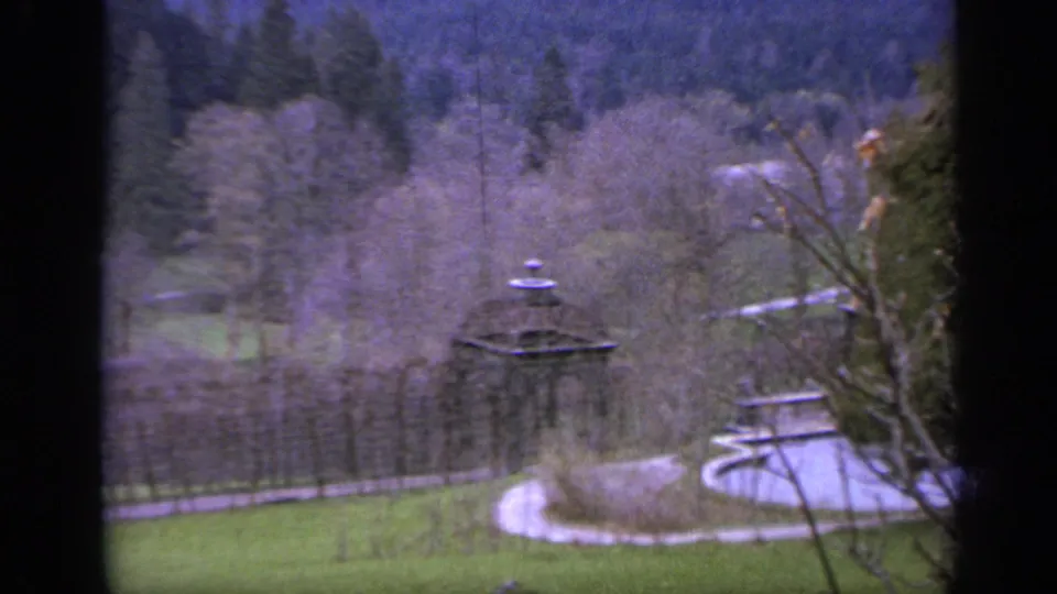 1962 Italy 8mm view of estate grounds with a fountain, winding garden path, bare trees, and forested mountain slopes beyond.