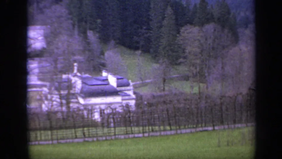 1962 Italy home-movie frame showing a large mansion near formal garden paths, lawns, bare trees, and wooded mountain terrain.