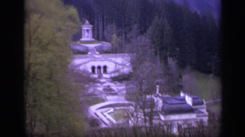 1962 Italy 8mm frame of a formal estate garden, fountain structure, terraced paths, mansion roofline, and dense trees.