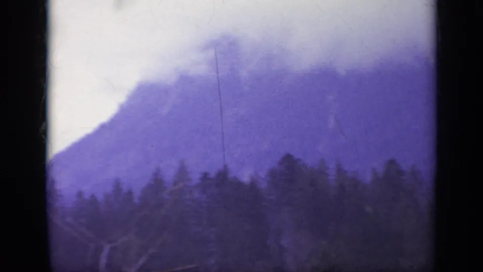 1962 Italy 8mm frame showing a dark treeline, broad hazy mountain slope, pale sky, and aged film scratches.