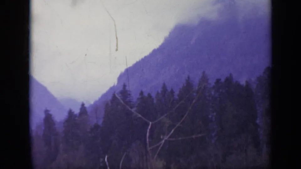 1962 Italy 8mm frame showing a hazy mountain ridge above evergreen trees, with purple-blue film fading and black frame edges.