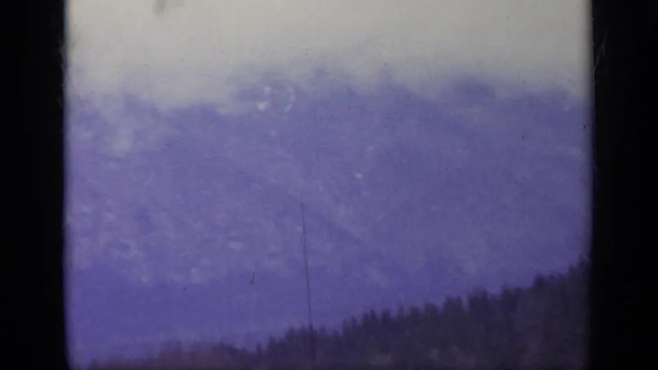 Faded 1962 Italy 8mm frame of a misty mountain face, with treetops at the bottom and a cool purple cast from aged film.
