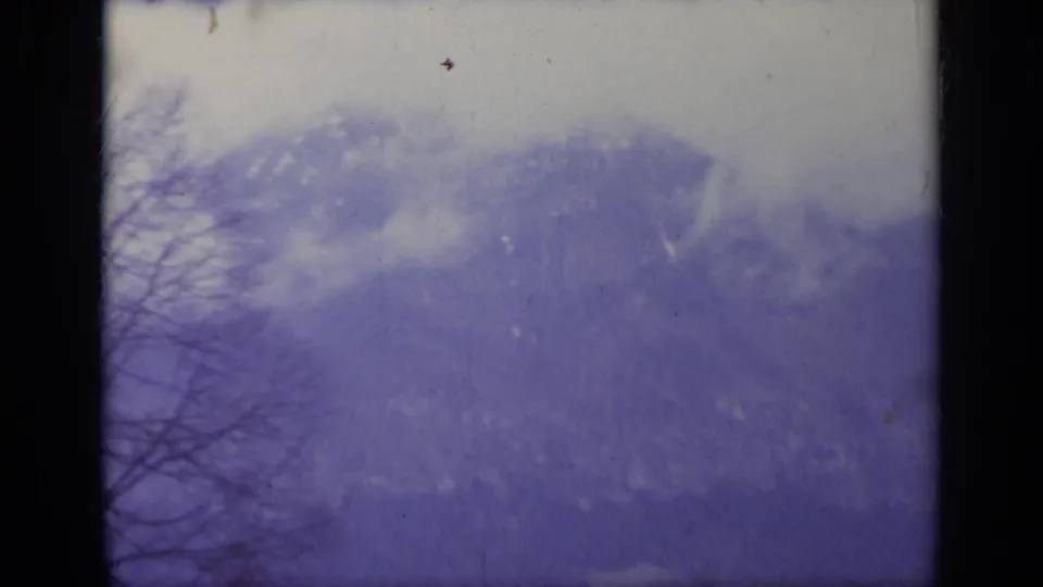 1962 Italy 8mm frame showing a fog-softened mountain ridge, bare branches, and aged film texture with a purple-blue shift.