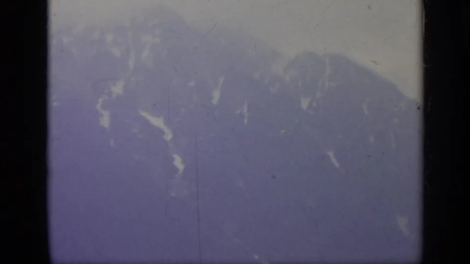 1962 Italy travel frame showing a distant snow-marked mountain ridge beneath a pale, hazy sky in soft 8mm color.
