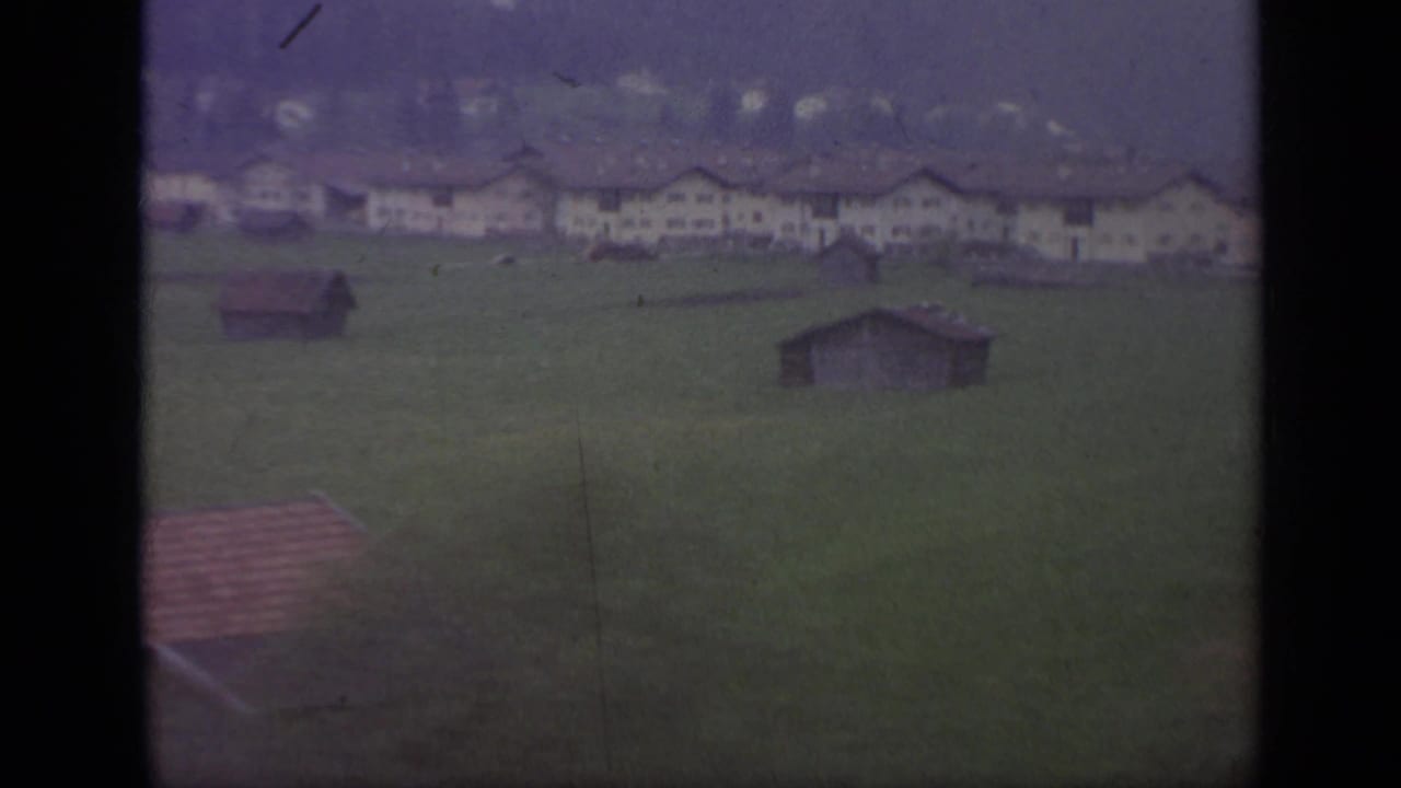 Green field with farm buildings and distant hills seen through a train window.