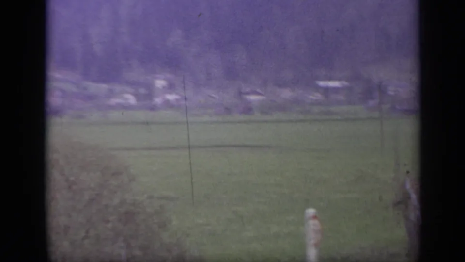 Rural Italian landscape from a train window, with open fields, distant buildings, hills, and a pale roadside post near the lower edge.