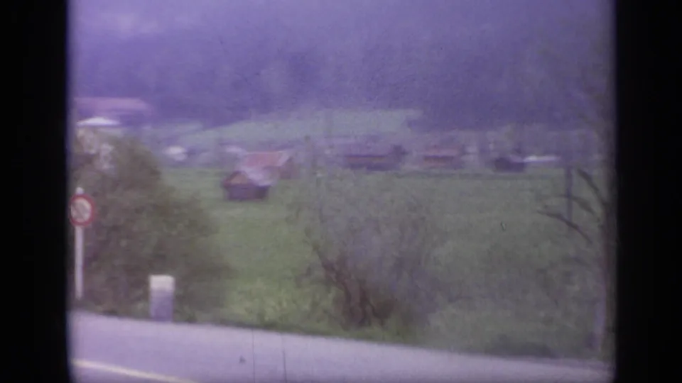 1962 Italy travel view with a roadside verge, circular sign, trees, green fields, and farm buildings blurred by train motion.
