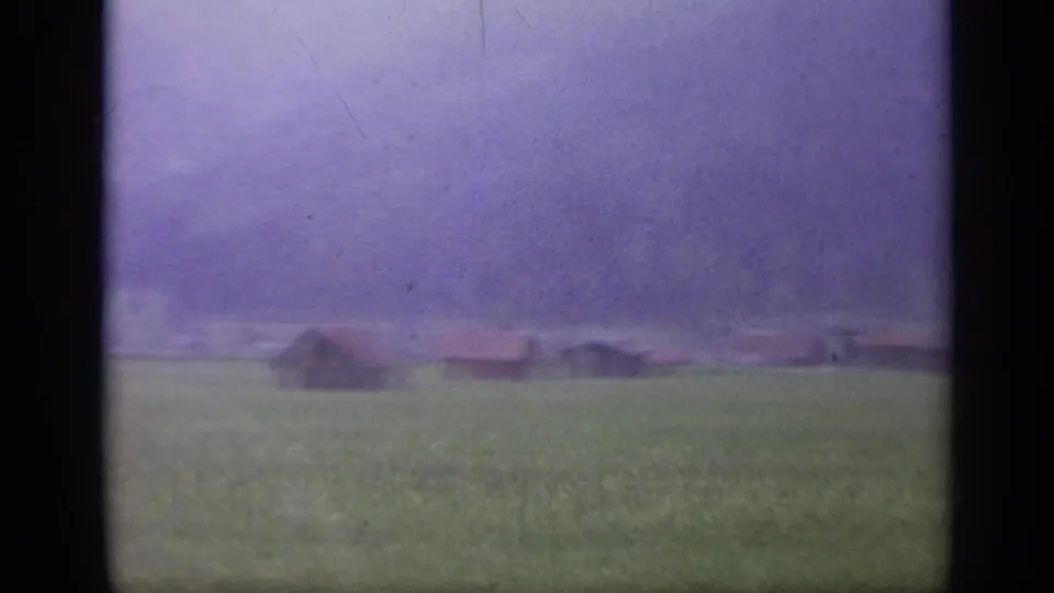 Rural Italy landscape from 1962 8mm footage, showing a green field, low farm structures, distant hills, and purple color fading.