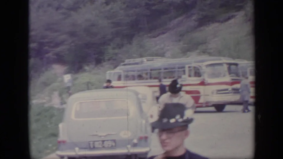 1962 Italy roadside stop with classic cars, red-and-white tour buses, and travelers in hats and coats near the road.