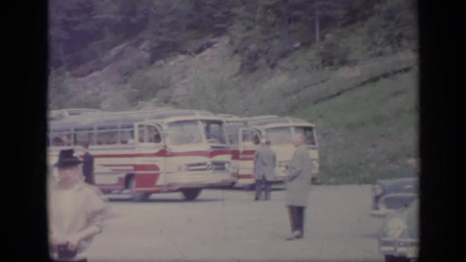Roadside view in 1962 Italy showing a red-and-white tour bus, several people, classic cars, and a grassy mountain slope.