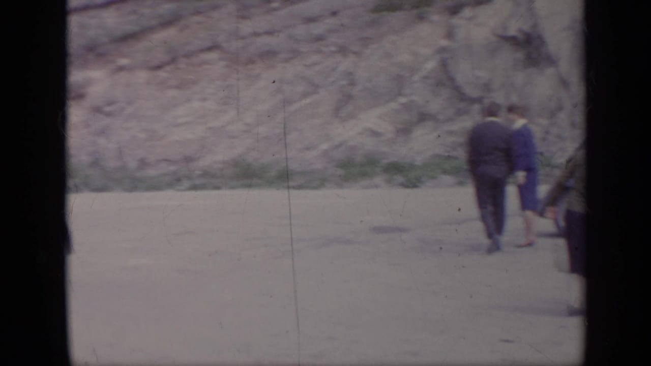People walk away along a roadside near parked cars and a rocky hillside.