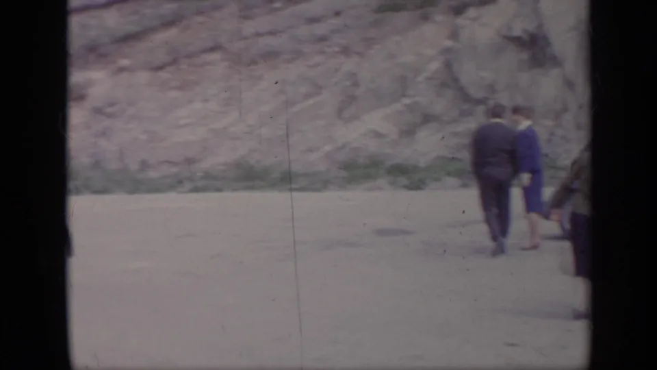 Travelers in period clothing walk along an Italian mountain roadside near parked cars in this 1962 home-movie frame.