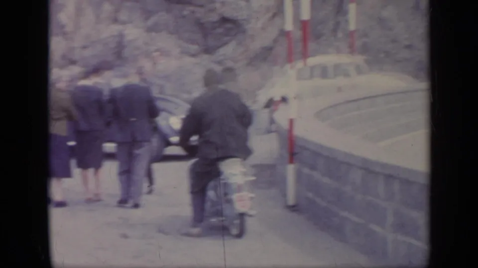 A small group stands near classic cars while a motorcyclist passes by at a 1962 Italian mountain roadside stop.