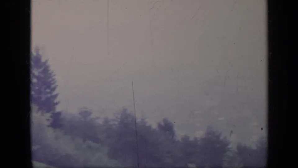 1962 Italy 8mm frame showing a hazy mountain landscape seen through dark vertical window bars, with treetops below.