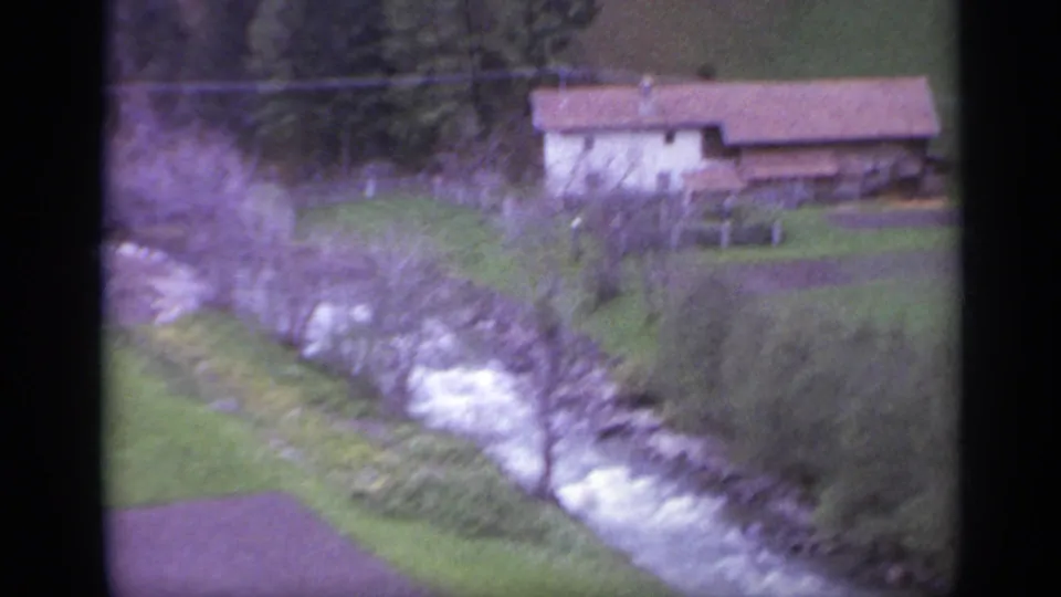 1962 Italian countryside frame showing a rocky stream, grassy banks, and a distant rural house with a reddish roof.