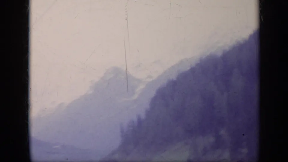 Hazy 1962 Italian alpine view with layered mountain ridges, dark fir-covered slopes, and a washed pale sky.