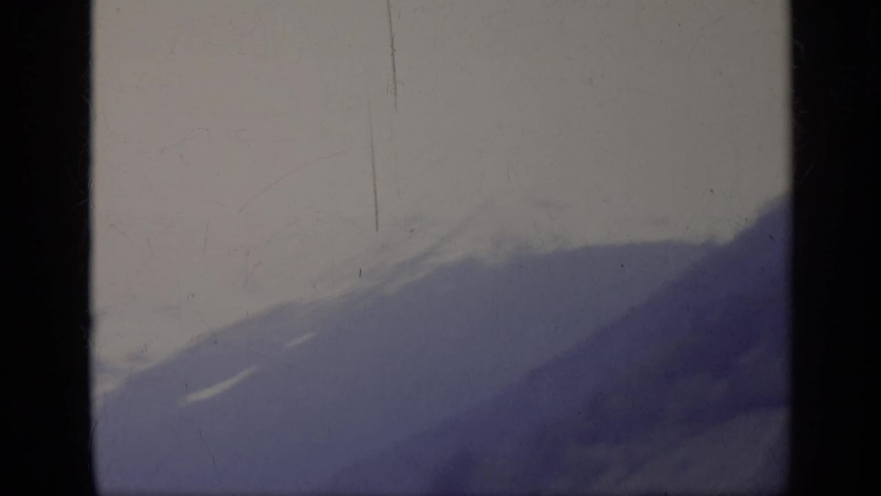 1962 8mm home-movie view in Italy of a hazy mountain slope and pale sky, framed by dark film edges.