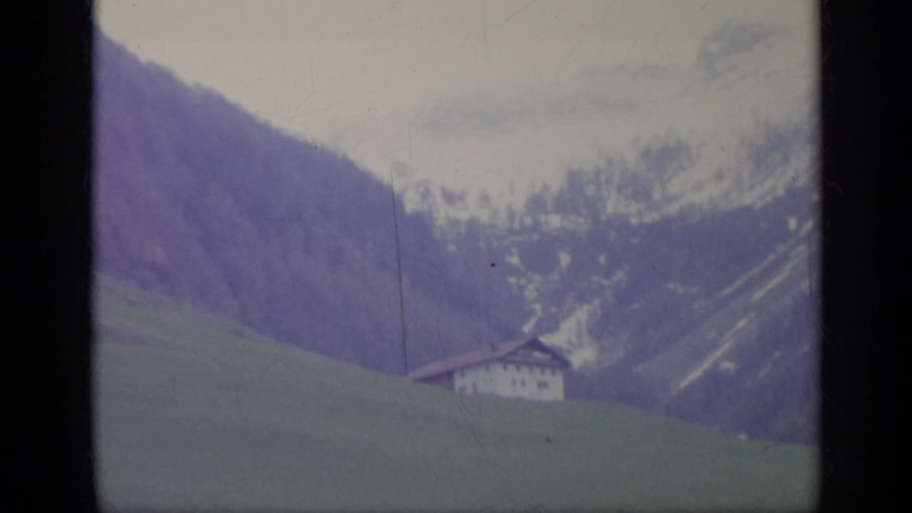 1962 Italian mountain landscape with a green slope, distant traditional house, and snow-marked ridges in soft 8mm color.