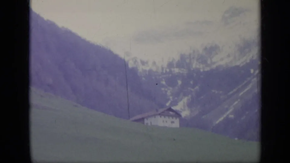 1962 Italian mountain landscape with a green slope, distant traditional house, and snow-marked ridges in soft 8mm color.