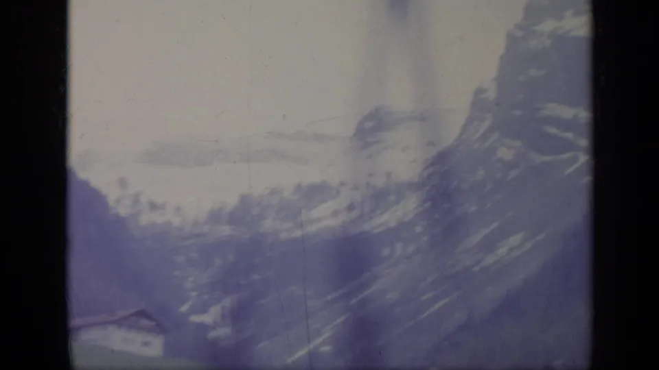 Vintage 8mm frame from Italy showing snow-streaked mountain terrain and the edge of a small alpine house.