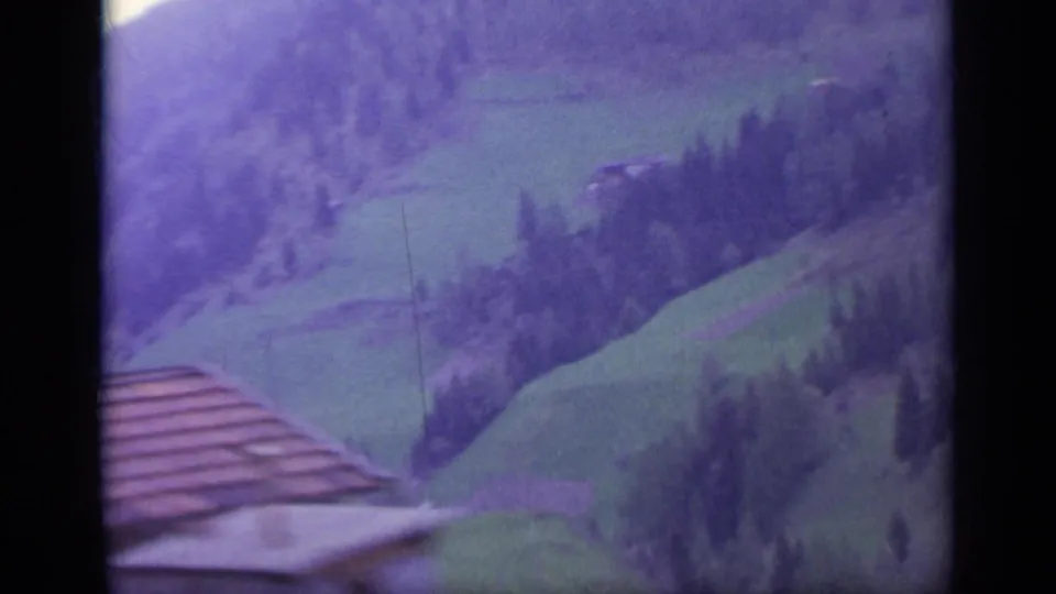 1962 Italy home-movie view over green valley fields and wooded mountain slopes, with a roofline at the lower left.
