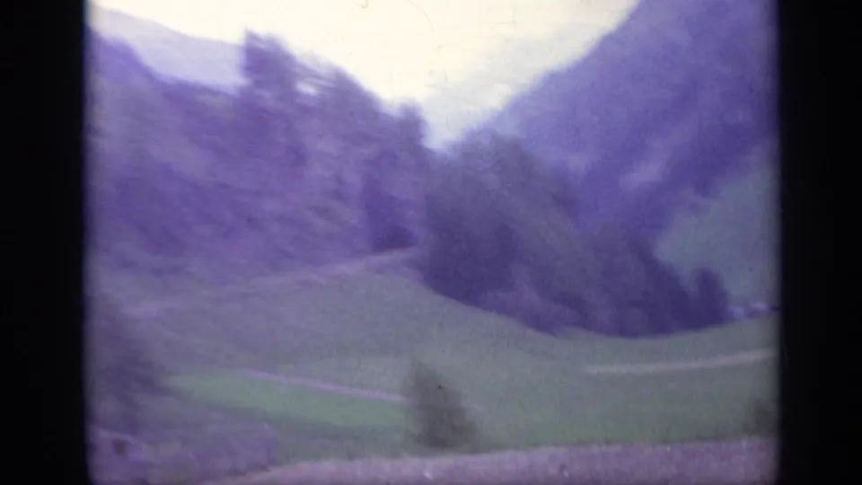Purple-faded 1962 Italy mountain valley view with green fields, wooded ridges, and a pale sky seen from a moving travel route.