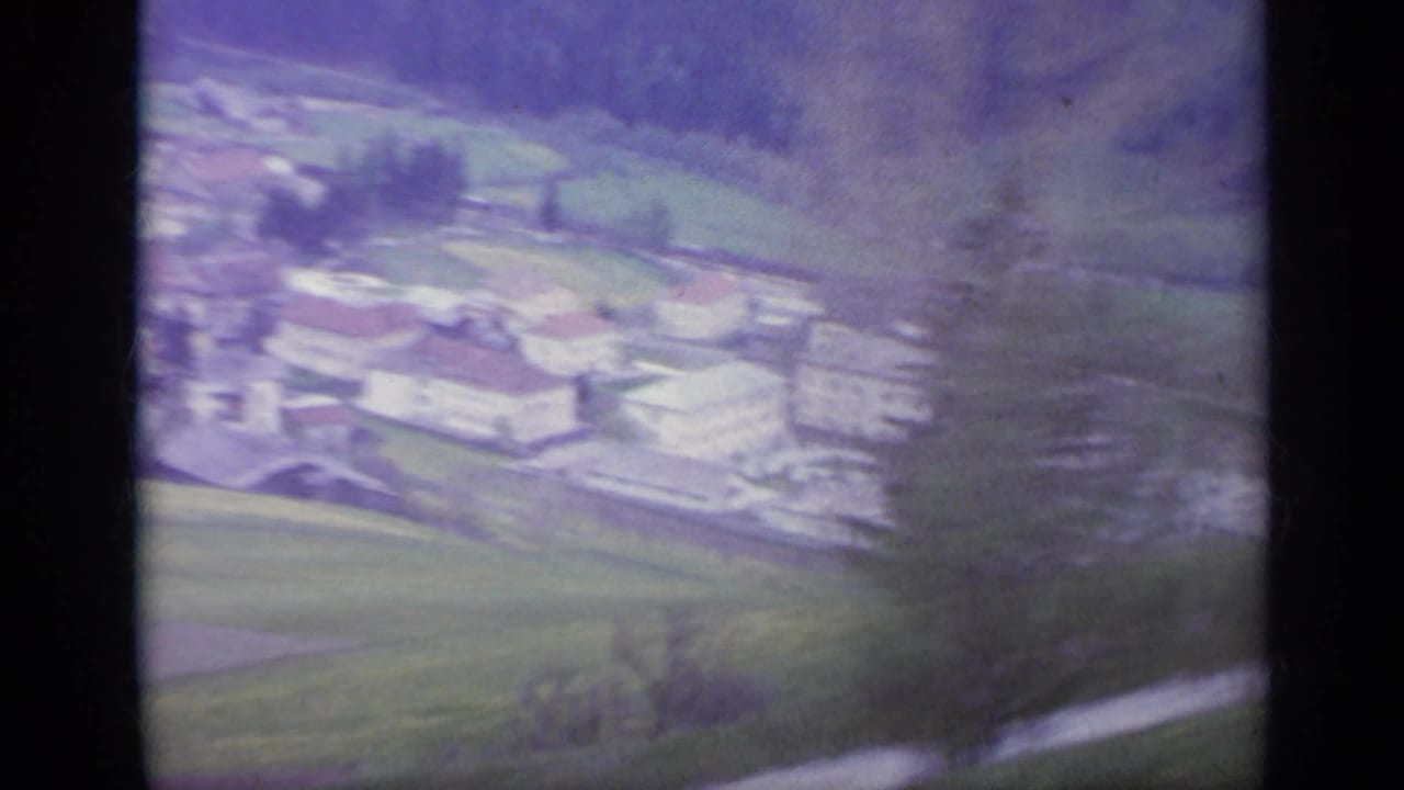 1962 Italy 8mm frame showing a soft, blurred view of an Alpine village, fields, and wooded hillside from a moving train.
