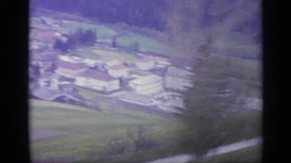 1962 Italy 8mm frame showing a soft, blurred view of an Alpine village, fields, and wooded hillside from a moving train.
