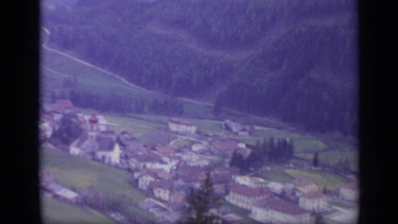 1962 Italy 8mm frame with a village, fields, tree lines, and dark forested mountain slope seen during the train journey.