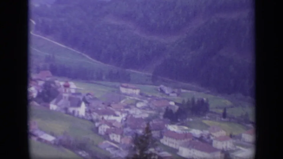 1962 Italy 8mm frame with a village, fields, tree lines, and dark forested mountain slope seen during the train journey.