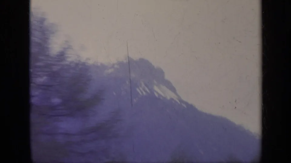 1962 Italy 8mm train-window view of a snow-capped alpine peak, trees, pale sky, and heavy motion blur.