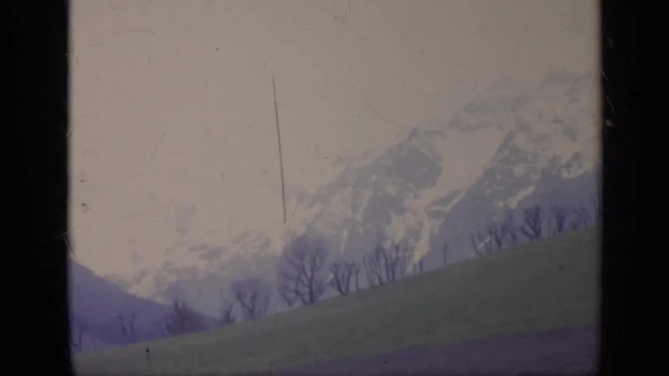 Italy 1962 8mm frame showing a soft, hazy mountain range beyond a rolling field and leafless roadside trees.