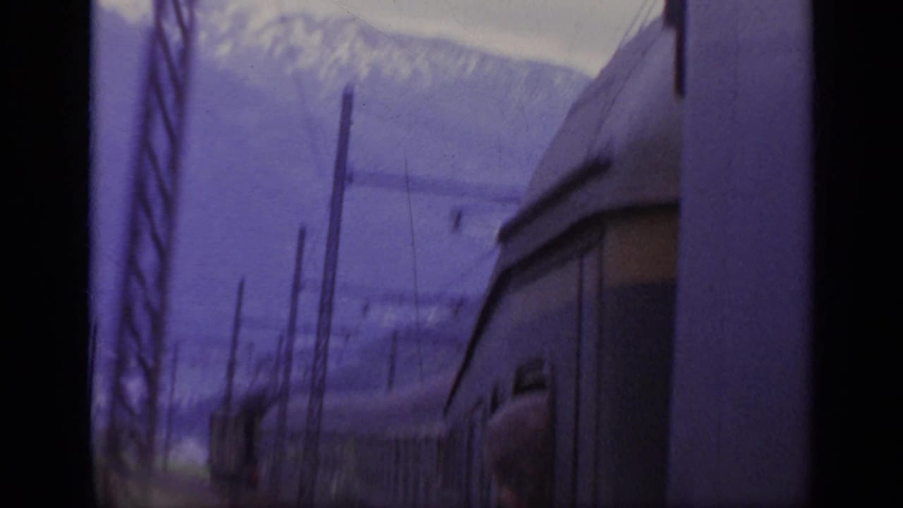 1962 Italy 8mm frame from a train, showing snowy alpine mountains, rail poles, overhead lines, and the side of a passing carriage.