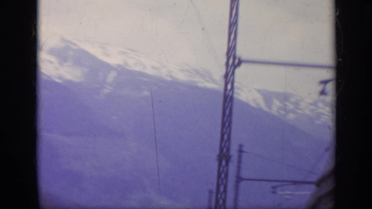 1962 Italy train-window view of a snow-covered mountain slope with overhead rail equipment and dark window framing.