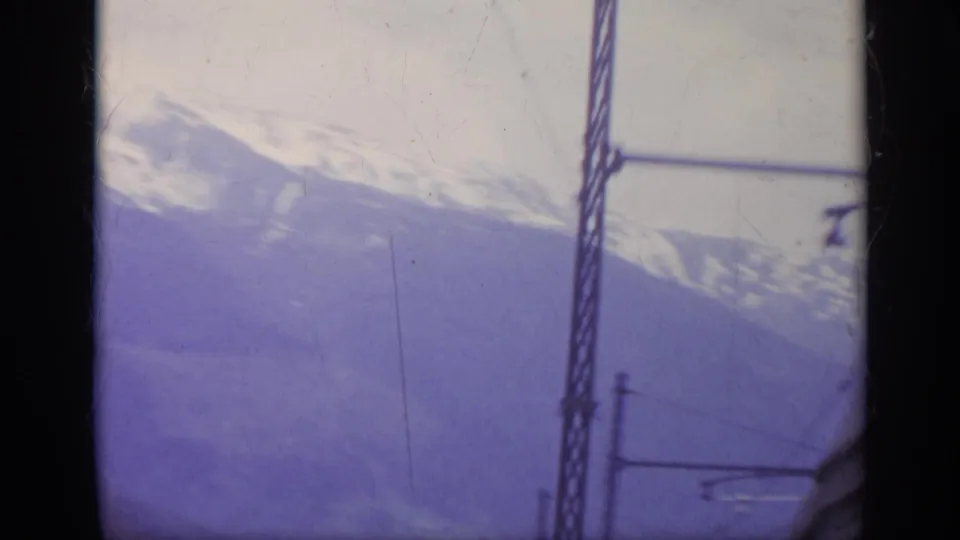 1962 Italy train-window view of a snow-covered mountain slope with overhead rail equipment and dark window framing.