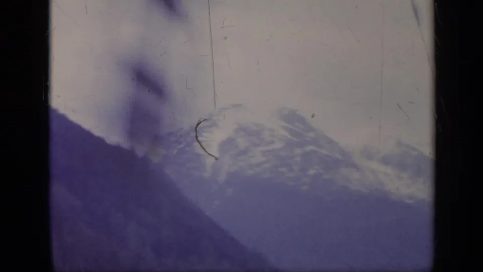 Soft, blurred 8mm train-view frame in Italy, with a snowy mountain slope, pale sky, and passing dark shapes near the window.