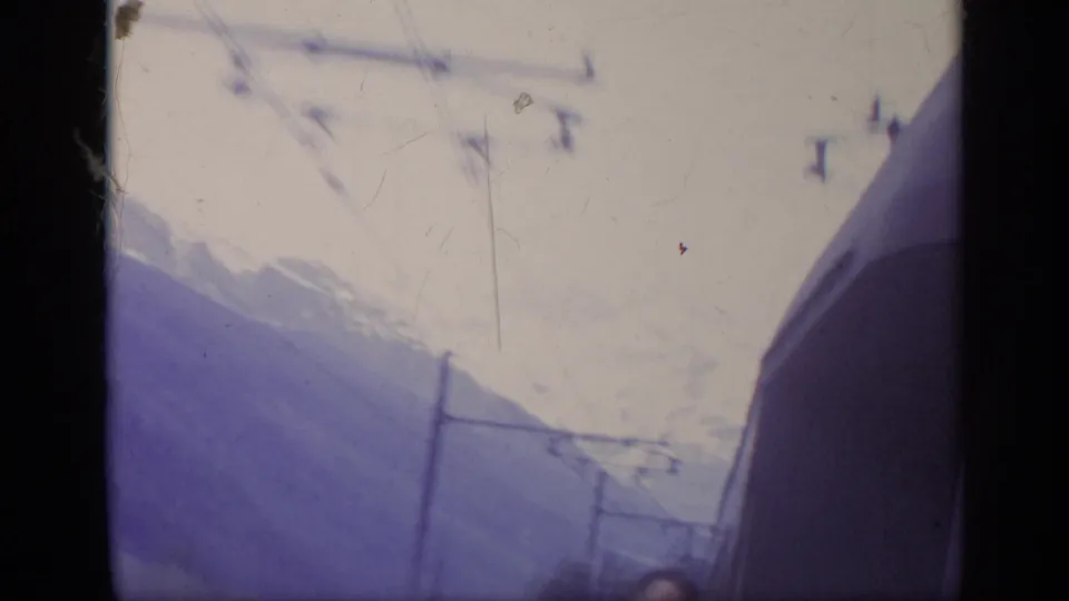 1962 Italy 8mm frame showing a passenger near the train window, with snow-covered mountains, rail poles, and overhead lines outside.
