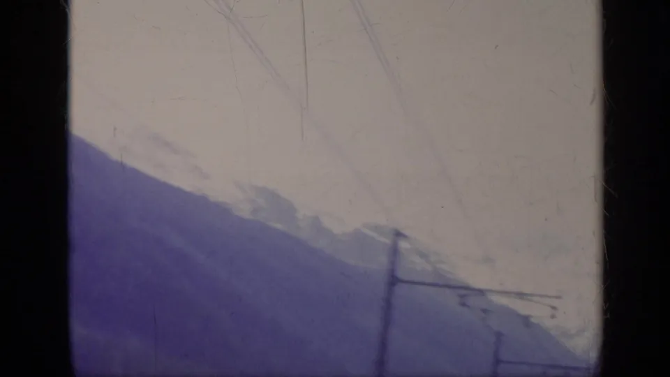 1962 Italy 8mm travel frame showing snow-capped mountains, valley slopes, and blurred overhead wires from a moving vehicle.