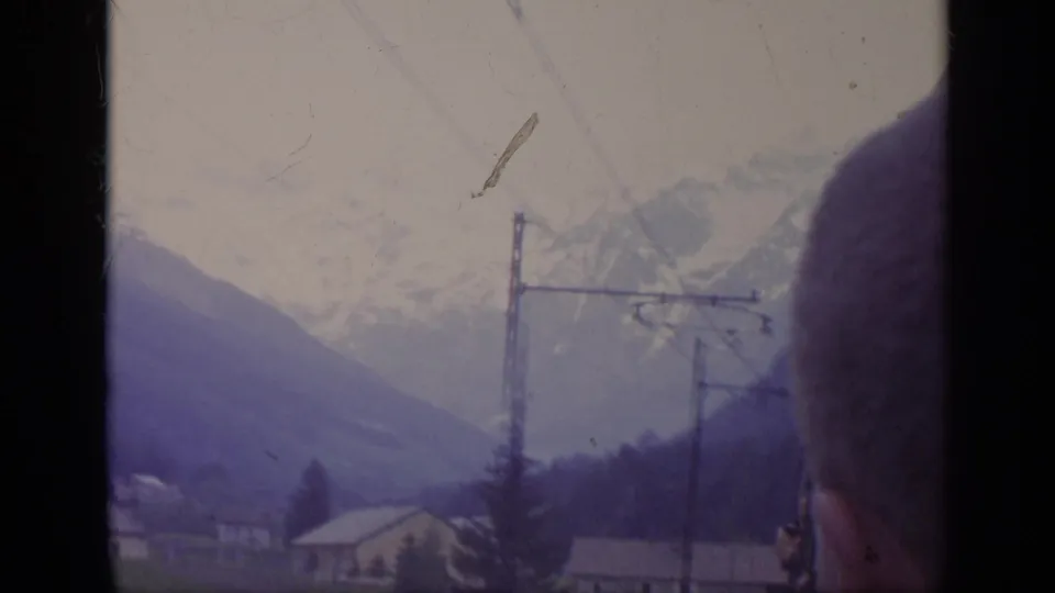 1962 Italy travel view from a moving vehicle, with alpine peaks, utility poles, valley buildings, and a partial passenger shape at right.