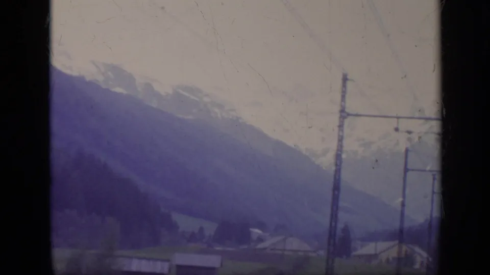 1962 Italy 8mm frame with alpine mountains, valley houses, wires, and tall poles blurred by vehicle movement.