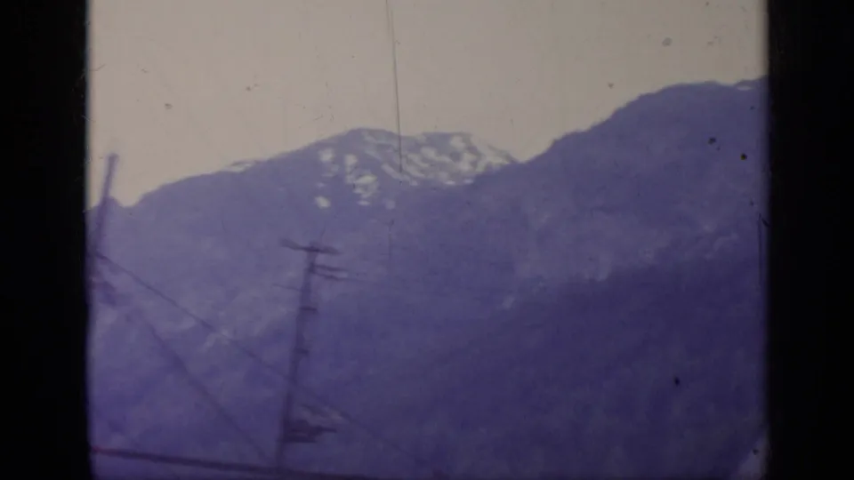 1962 Italy alpine travel frame showing a mountain slope, snowy ridge, utility pole, and window-framed motion blur.